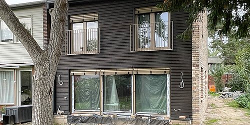 Foto, Zweistöckiges Neubau-Strohhaus mit dunkler Holzfassade, großen Fenstern und kleinem Balkon, umgeben von Gras und Bäumen