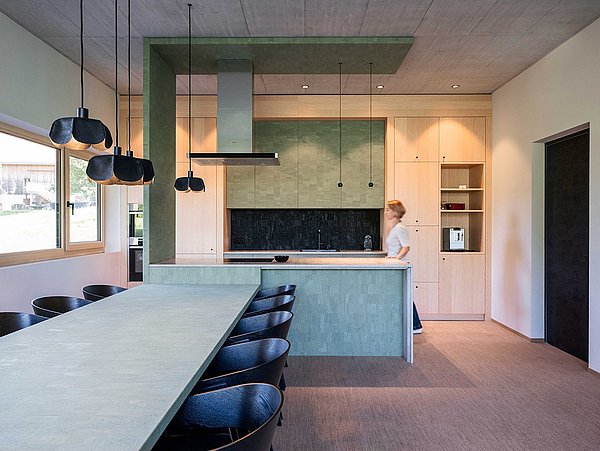 Foto, moderner Küchenraum mit langem Esstisch, schwarzen Stühlen, grüner Kücheninsel und einer Person im Hintergrund