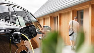 Foto, Elektroauto mit Ladekabel an Ladestation im Vordergrund und unscharf gehender Mann vor Holzhaus im Hintergrund.