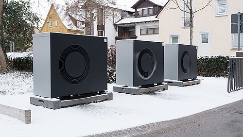 Foto, drei graue Wärmepumpen stehen auf Paletten auf schneebedecktem Boden vor Wohnhäusern.