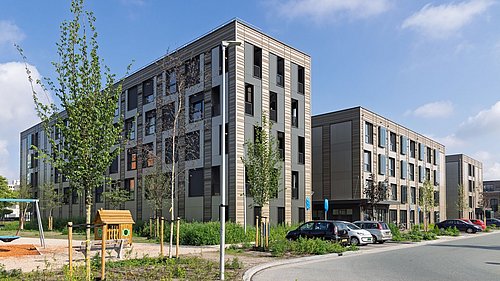 Foto, Modernes Wohngebäude mit hellen Fassaden, mehreren Fenstern und Parkplätzen, davor Spielplatz mit Schaukel und Sandkasten.