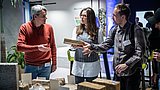 Foto, drei Personen stehen an einem Tisch mit rechteckigen, braunen Proben, eine Person hält eine Probe in der Hand, im Hintergrund Pflanzen und blaue Beleuchtung.