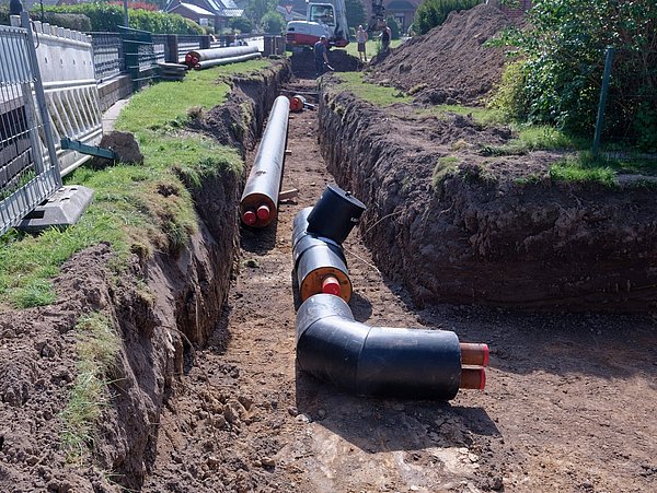 Foto, Verlegte große schwarze Kunststoffrohre mit roten Innenrohren in einem offenen Graben neben einem Zaun auf einer Baustelle