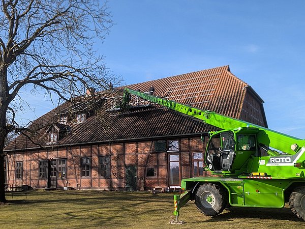 Foto, Grüner Teleskoplader mit der Aufschrift 'ROTO' hebt Arbeiter auf das Dach eines großen Backsteingebäudes mit roten Dachziegeln