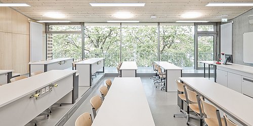 Foto, Klassenzimmer mit zwei Tischreihen und Lehrerpult. Die Wände sind teilweise aus Beton, teilweise aus Holz. Im Hintergrund befindet sich eine große Fensterfront.