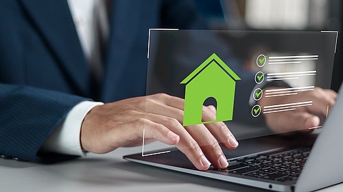 Foto, Hände tippen auf Laptop-Tastatur vor halbtransparentem Bildschirm mit grünem Haussymbol und vier grünen Häkchen.