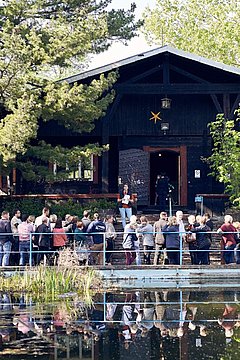 Foto, Gruppe von Menschen hört einem Vortrag vor einer dunklen Blockhütte umgeben von Bäumen zu, im Vordergrund ein Teich mit Spiegelung.