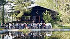 Gruppe von Menschen hört einem Vortrag vor einer dunklen Blockhütte umgeben von Bäumen zu, im Vordergrund ein Teich mit Spiegelung.