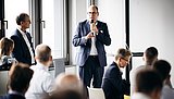 Foto, Mann im Anzug hält Mikrofon und spricht vor sitzendem Publikum in einem hellen Konferenzraum
