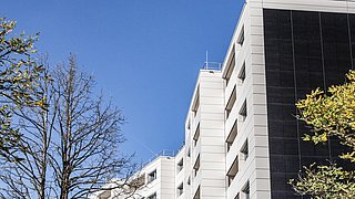 Foto, moderne Gebäudefassade mit großen Fenstern und Sonnenkollektoren, eingerahmt von kahlen und grünen Bäumen vor einem klaren blauen Himmel.