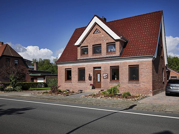 Foto, Zweistöckiges rotes Backsteinhaus mit weißem Giebel und roten Dachziegeln an einer Straße bei klarem Himmel.