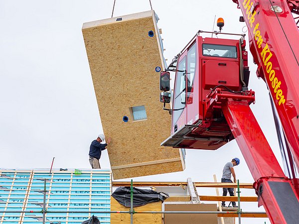 Foto, zwei Bauarbeiter auf einem Gerüst montieren mit einem roten Kran ein großes vorgefertigtes Wandpaneel aus Holzwerkstoff an einem Dach.