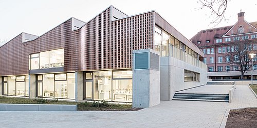Foto, Aufnahme des neuen Schulgebäudes aus Holz und Beton. Im Hintergrund befindet sich der alte Klinkerbau des Schulgeländes. 