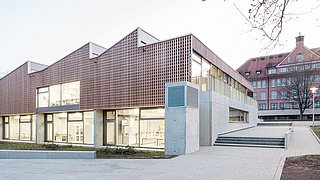 Foto, Aufnahme des neuen Schulgebäudes aus Holz und Beton. Im Hintergrund befindet sich der alte Klinkerbau des Schulgeländes. 