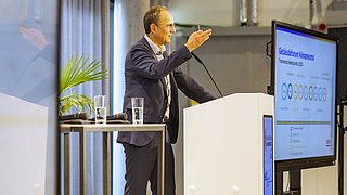 Foto, ein Mann im Anzug gestikuliert, während er auf einem Podium vorträgt. Auf einem großen Bildschirm werden in einer modernen Konferenzumgebung die Themen angezeigt.