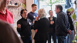 Foto, Gruppe von Menschen in einem hellen Raum im Gespräch, einige halten Getränke in der Hand.