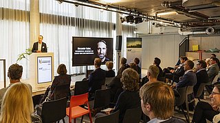 Foto, ein Redner präsentiert auf der Bühne vor einem Publikum in einem modernen Konferenzraum mit Vorhängen und Bildschirmen, auf denen das Thema angezeigt wird.