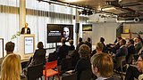 Foto, ein Redner präsentiert auf der Bühne vor einem Publikum in einem modernen Konferenzraum mit Vorhängen und Bildschirmen, auf denen das Thema angezeigt wird.