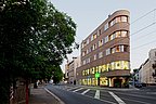 Foto, Ansicht von der Straße eines mehrstöckigen, spitz zulaufenden Gebäudes mit Holzfassade. Links im Bild steht ein Baum.