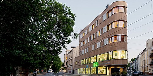 Foto, Ansicht von der Straße eines mehrstöckigen, spitz zulaufenden Gebäudes mit Holzfassade. Links im Bild steht ein Baum.