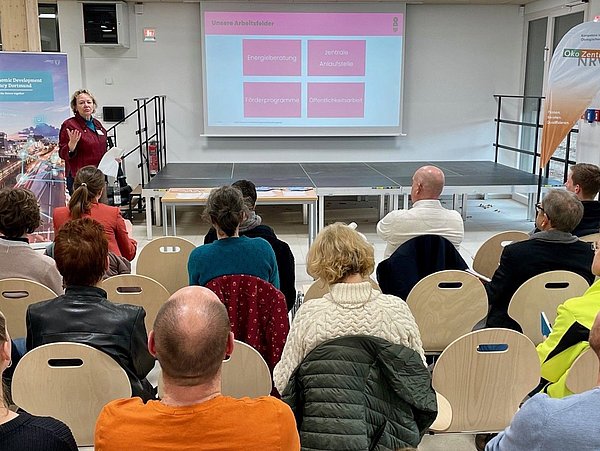Foto, Eine Frau steht vor einer Gruppe sitzender Menschen und präsentiert Folien auf einer Leinwand in einem Veranstaltungsraum.