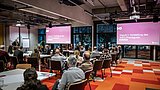Foto, Konferenzraum mit mehreren Personen, die auf zwei große Bildschirme mit Präsentationen schauen.