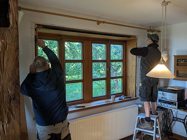 Foto, Zwei Männer in Arbeitskleidung arbeiten an Fensterlaibung mit Holzfenster in einem Raum mit sichtbaren Holzbalken und einem Heizkörper unter dem Fenster