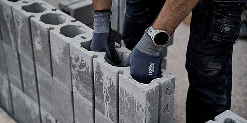Foto, Hand mit Schutzhandschuh greift in eine Öffnung eines Betonsteins, im Hintergrund weitere Steine und eine Person in Arbeitskleidung.