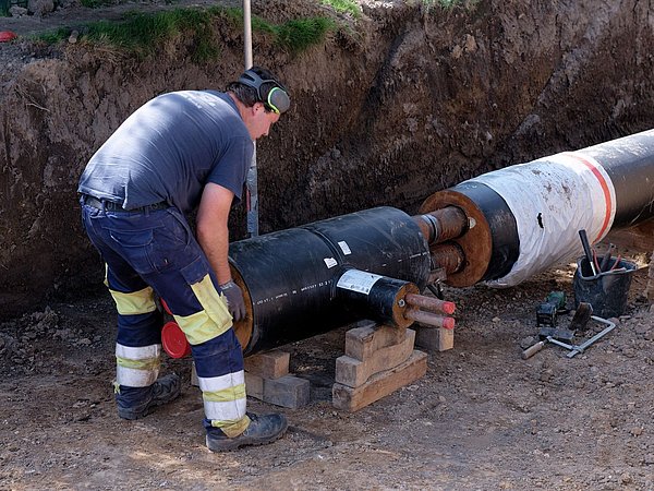 Foto, Arbeiter montiert isolierte Rohrleitung in einer Baugrube, trägt Schutzkleidung und Gehörschutz