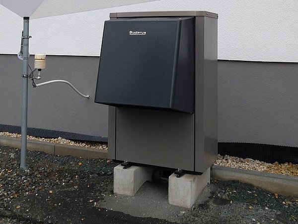 Foto, Außeneinheit einer Wärmepumpe mit schwarzer Abdeckung an einer weißen Hauswand