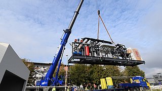 Ein blauer Kran hebt eine große schwarze Energiezentrale mit Rohren und einem roten Zylinder, mehrere Personen beobachten die Anlieferung im Freien bei blauem Himmel.