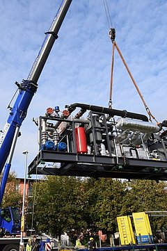 Foto, ein blauer Kran hebt eine große schwarze Energiezentrale mit Rohren und einem roten Zylinder, mehrere Personen beobachten die Anlieferung im Freien bei blauem Himmel.