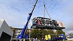 Ein blauer Kran hebt eine große schwarze Energiezentrale mit Rohren und einem roten Zylinder, mehrere Personen beobachten die Anlieferung im Freien bei blauem Himmel.