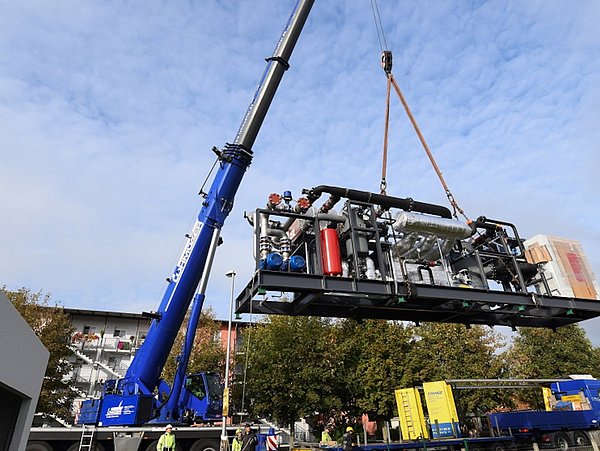 Ein blauer Kran hebt eine große schwarze Energiezentrale mit Rohren und einem roten Zylinder, mehrere Personen beobachten die Anlieferung im Freien bei blauem Himmel.