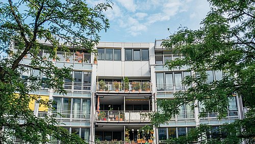 Foto, Mehrstöckiges Wohngebäude mit Balkonen, auf denen Pflanzen und Möbel stehen, umgeben von grünen Bäumen