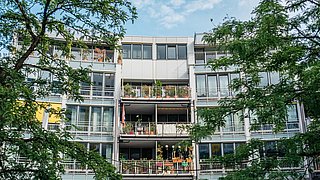 Foto, Mehrstöckiges Wohngebäude mit Balkonen, auf denen Pflanzen und Möbel stehen, umgeben von grünen Bäumen