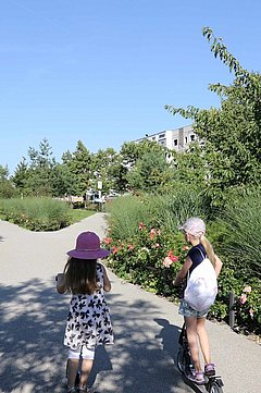 Foto, Zwei Kinder mit Sommerkleidung und Hüten fahren auf einem breiten Weg durch eine grüne Wohnsiedlung mit modernen mehrstöckigen Gebäuden, die Wege kreuzen sich und bilden eine Achse
