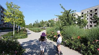 Foto, Zwei Kinder mit Sommerkleidung und Hüten fahren auf einem breiten Weg durch eine grüne Wohnsiedlung mit modernen mehrstöckigen Gebäuden, die Wege kreuzen sich und bilden eine Achse