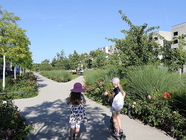 Foto, Zwei Kinder mit Sommerkleidung und Hüten fahren auf einem breiten Weg durch eine grüne Wohnsiedlung mit modernen mehrstöckigen Gebäuden, die Wege kreuzen sich und bilden eine Achse