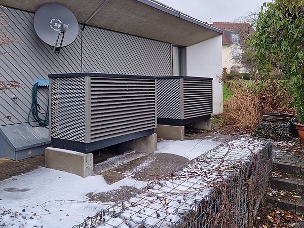 Foto, Zwei mit Metallgittern verkleidete Außengeräte auf Betonplatten vor einem Haus mit grauer Fassade und Satellitenschüssel im Winter, es liegt Schnee