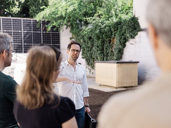 Mehrere Personen stehen im Außenbereich mit Bäumen und Mauer vor einer Wand mit einer Photovoltaikanlage und einem Absorberkasten.