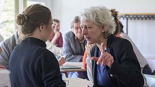 Foto, zwei Personen im Gespräch, eine zeigt mit dem Zeigefinger nach oben, im Hintergrund weitere Personen an Tischen in einem hellen Raum