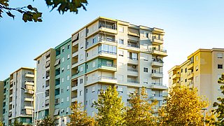 Foto, mehrstöckiges Wohngebäude mit Balkonen und Markisen in Pastellfarben unter klarem Himmel, umgeben von Bäumen.