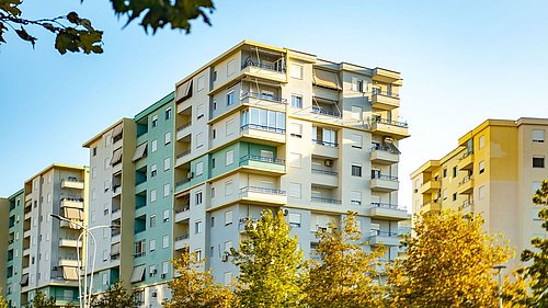 Foto, mehrstöckiges Wohngebäude mit Balkonen und Markisen in Pastellfarben unter klarem Himmel, umgeben von Bäumen.
