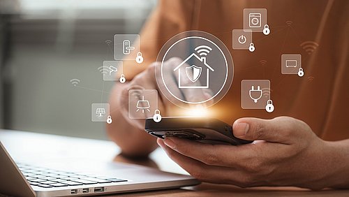 Foto, Hand hält Smartphone vor Laptop mit schwebenden Symbolen für Smart-Home-Sicherheit und vernetzte Geräte.