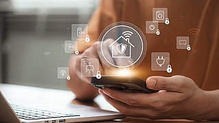 Foto, Hand hält Smartphone vor Laptop mit schwebenden Symbolen für Smart-Home-Sicherheit und vernetzte Geräte.