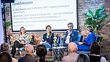 Foto, Podiumsdiskussion mit vier Teilnehmenden auf Stühlen vor Publikum, Hintergrund mit Backsteinwand und Pflanzen, Präsentationsfolie mit Namen und Titeln der Sprecher