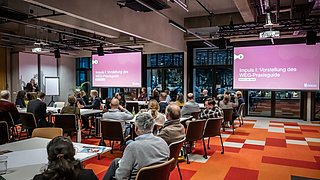 Foto, Konferenzraum mit mehreren Personen, die auf zwei große Bildschirme mit Präsentationen schauen.