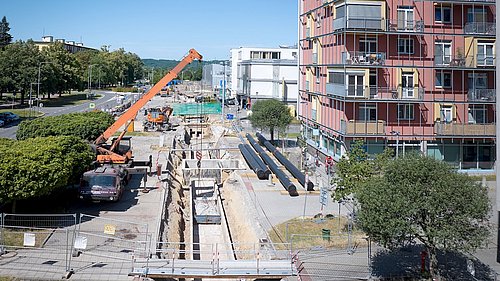 Foto, Baustelle mit tiefem Graben, mehreren großen schwarzen Rohren, einem orangen Kran und einem mehrstöckigen Wohngebäude im Hintergrund.
