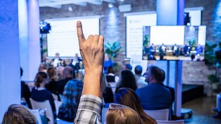 Foto, Hand einer Person hebt sich in einem Publikum während einer Konferenz mit Podiumsdiskussion im Hintergrund